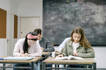 Students sitting in a classroom with a blackboard behind them. The students are looking at textbooks on their desks and seem to be studying.