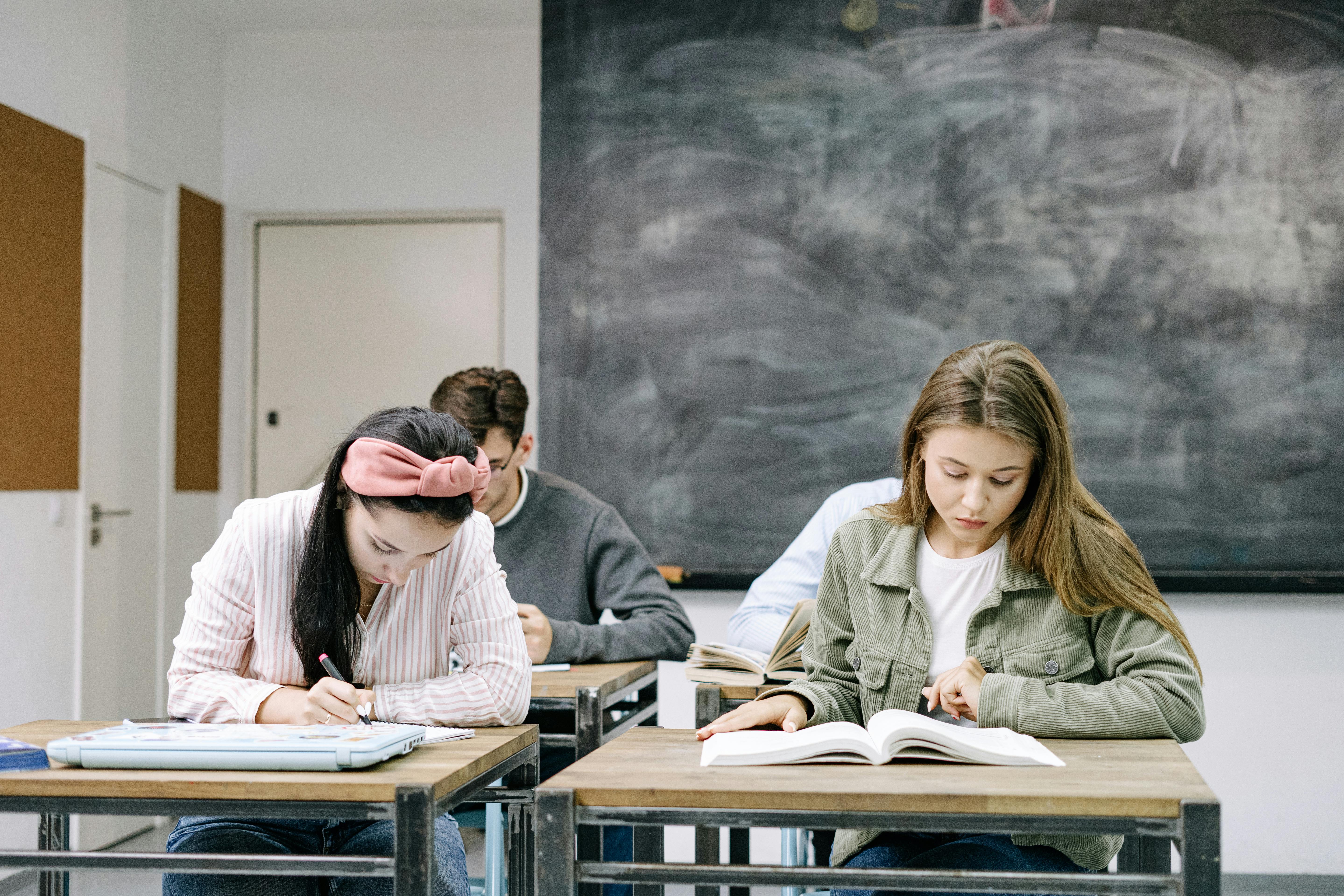 Students sitting in a classroom with a blackboard behind them. The students are looking at textbooks on their desks and seem to be studying.